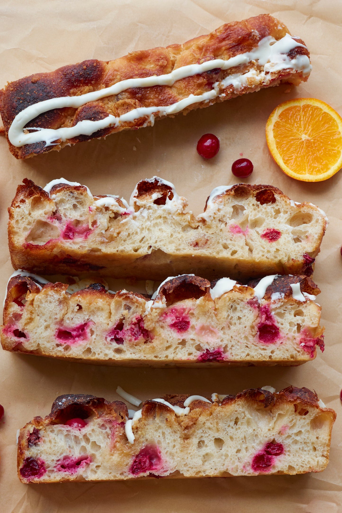 Sliced cranberry bread with orange glaze on parchment paper with oranges and cranberries around it. 